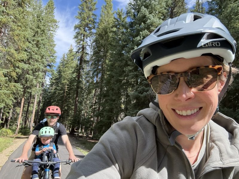 The founder smiling on a paved forest path, with her husband and their son, in a front-mounted child seat, riding right behind her